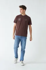 Remera de hombre color marrón chocolate, con corte regular, escote redondo y mangas cortas. Presenta un diseño estampado pequeño en el pecho con motivos gráficos y texto.