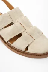 Sandalia tipo cangrejera de cuero color arena, con diseño de tiras anchas entrelazadas, talón descubierto y cierre mediante hebilla metálica lateral. Posee suela plana de goma color marrón con textura antideslizante.