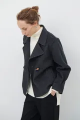 Chaqueta de paño color gris oscuro, con solapas amplias y cierre frontal mediante un alamar único. Presenta bolsillos laterales y mangas largas con puños rectos.