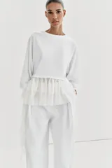 Camiseta de algodón con cuello redondo y manga corta. Presenta un bajo asimétrico confeccionado en un tejido ligero con volantes y una tira lateral decorativa con volantes.