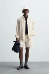 Sobrecamisa de hombre de corte relajado, color beige claro con finas rayas verticales en tono más oscuro. Presenta cuello bowling, manga larga con puño abotonado, bolsillos de parche en el pecho y en la cadera, y aberturas laterales en el bajo.