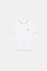 Camiseta negra de algodón, corte regular, con cuello redondo ribeteado en blanco y un bolsillo frontal con un broche metálico decorativo dorado.