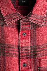 Camisa de sarga de algodón con diseño a cuadros, cuello inglés, bolsillo en el pecho y cierre frontal de botones. Presenta mangas largas con puños abotonados y bajo redondeado.