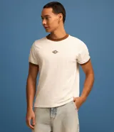 Remera estilo baby tee de color blanco, confeccionada en algodón, con cuello redondo y ribetes en color marrón en cuello y puños. Cuenta con un pequeño tag gomosa con el logotipo "Lucky" en el centro del pecho.