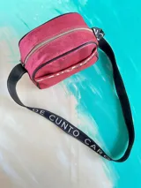 Bandolera Kaira de la línea Ottenta de Carolina De Cunto, en color fucsia metalizado. Cuenta con un diseño compacto, bolsillo exterior con broche imantado y detalle pintado a mano, cierre superior y una correa larga regulable con el logo de la marca estampado.