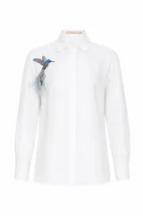 Camisa de lino celeste con cuello camisero, manga larga y cierre frontal oculto con botones. Incluye un broche desmontable bordado con forma de colibrí y plumas en el pecho.