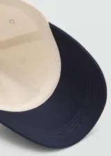 Gorra Archive de color beige con visera y botón superior azul marino. Cuenta con mensaje bordado en el lateral y cierre regulable con hebilla metálica.