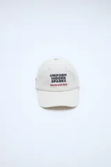 Gorra gris con visera curva, bordado frontal con texto en color beige y detalle bordado lateral.