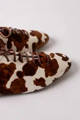 Zapato tipo flat de estilo acordonado, confeccionado en cuero con pelo con estampado de manchas tipo vaca en tonos marrón y blanco.
