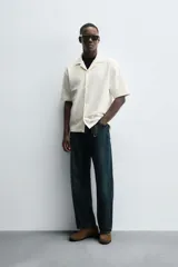 Camisa de manga corta con cuello bowling y calce relaxed fit, confeccionada en tejido seersucker elástico con textura. Presenta un diseño a rayas verticales en tonos oscuros y cierre frontal con botones.