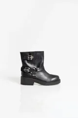 Botas de caña baja confeccionadas en cuero sintético negro, con suela plana y taco bajo. Presentan detalles de correas con hebillas metálicas y una argolla decorativa en el tobillo.