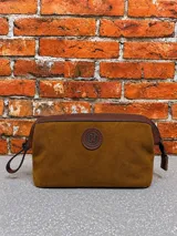 Necessaire de diseño rectangular confeccionado en gamuza color tostado, con detalles de cuero marrón en los bordes superiores y cierre metálico. Presenta un aplique circular de cuero con logo grabado en el centro.