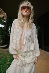 Blusa de escote en pico y manga larga con puños elásticos. Presenta un diseño semitransparente con bordados florales a tono, cintura elástica y cierre frontal mediante lazos con abalorios decorativos.