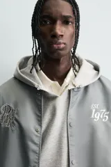 Cazadora estilo varsity o bomber, cuerpo principal gris oscuro y mangas color crema. Presenta cuello, puños y bajo en rib elástico con rayas grises y blancas. Tiene bordados en el pecho: un monograma estilizado a la izquierda y el texto "EST. 1975" a la derecha.