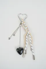 Llavero con mosquetón metálico en forma de corazón, cuerdas en tonos beige y blanco con detalles negros, cadenas colgantes con dijes de estrella y corazón, y un dije principal de corazón negro brillante.
