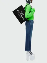 Bolso tipo tote de Marc Jacobs, confeccionado en lona resistente de color negro. Presenta el nombre del modelo estampado en blanco en la parte frontal y el logo de la marca debajo. Cuenta con doble asa superior para llevar al hombro o en la mano.
