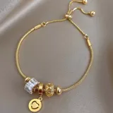 Pulsera dorada con cadena de eslabones y cierre corredizo ajustable. Incluye dijes colgantes: uno con forma de corazón y sonrisa, otro con forma de corazón calado, uno con forma de cilindro con incrustaciones de strass y una cuenta redonda lisa.
