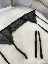Conjunto de lencería de encaje negro compuesto por un bralette triangular, una tanga y un portaligas integrado. El diseño presenta detalles de encaje floral, breteles regulables y tiras elásticas ajustables.