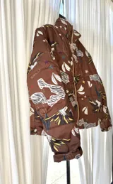 Campera puffer color marrón habano con estampado de loros, estrellas y elementos botánicos, cuello alto y cierre frontal.