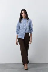 Camisa de rayas verticales con cuello mao, detalle de volados en el escote, bordados florales y recortes con calados. Posee mangas largas abullonadas con puños ajustados y cierre frontal con botones.