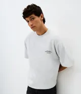 Remera gris melange de corte relaxed, con cuello redondo y mangas cortas. Presenta un diseño gráfico en la espalda con estampado y textura en alto relieve embossed que incluye la frase "Beyond Limits: Keep Going" y el texto "The Urban Supply Co. of N.Y.C.".
