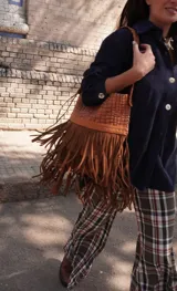 Cartera de hombro de cuero color tostado con diseño amplio, cuerpo superior texturizado y flecos largos colgantes en el frente. Cuenta con una correa trenzada para llevar al hombro.
