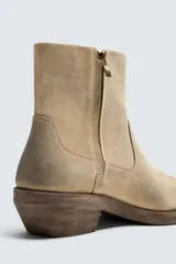 Bota cowboy de caña media, color beige claro con efecto desgastado. Fabricada en piel serraje engrasado, presenta horma cuadrada y tacón ancho de madera a contraste.