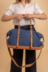Bolso de viaje amplio confeccionado en tela canvas azul con detalles, base, asas y correas en símil cuero tostado. Cuenta con correas ajustables y herrajes metálicos.