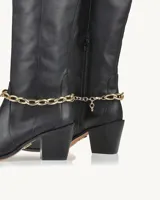 Bota alta de cuero negro con punta fina, taco geométrico y detalle de cadena dorada en el tobillo.