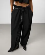Pantalón negro de corte ancho y tiro alto con diseño asimétrico. Presenta un cruce frontal con cinturón de tela a cuadros grises y negros, que deja ver un forro o panel interior de la misma tela a cuadros.