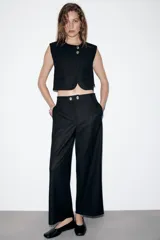 Pantalón negro de tiro medio con pierna ancha y cierre frontal de cremallera con dos botones dorados decorativos.