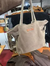 Bolso tipo tote de cuero descarne color visón, con diseño artesanal, flecos en la parte trasera y hebillas metálicas laterales. Cuenta con doble asa de hombro, doble bolsillo interno y tira porta llaves.
