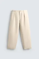 Pantalón relaxed fit color beige, confeccionado en tejido de algodón, con pliegues frontales, bolsillos delanteros y bolsillos traseros de vivo. Presenta un efecto lavado y cierre frontal con cremallera y botón.