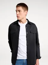 Camisa de sarga negra con cierre frontal de botones a presión, cuello clásico y dos bolsillos frontales con solapa.