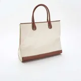 Bolso tipo tote de estructura rígida, confeccionado en lona color beige con detalles de cuero sintético en color marrón en la base, las asas y los bordes superiores.