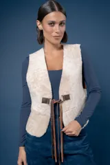 Chaleco de piel sintética en color beige, con cierre frontal mediante hebillas metálicas y tiras de cuero marrón que cuelgan como detalle decorativo.