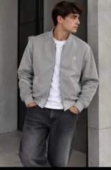 Campera bomber gris claro de paño fino, con cierre frontal metálico, bolsillos ribeteados y cuello, puños y ruedo de rib elástico. Presenta un pequeño bordado distintivo en el pecho.