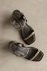 Sandalia de cuero color bronce metalizado, con punta cuadrada, taco ancho y bajo, y tiras finas adornadas con tachas esféricas plateadas.