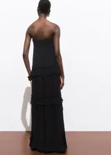 Vestido largo de fiesta color negro, con escote palabra de honor y diseño evasé. Presenta múltiples niveles con volantes decorativos y cierre lateral con cremallera.