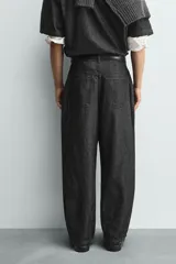 Pantalón de jeans de algodón ligero, color negro lavado, con corte balloon fit (holgado y ancho). Presenta pliegues frontales marcados en la cintura y cierre frontal de cremallera y botón.