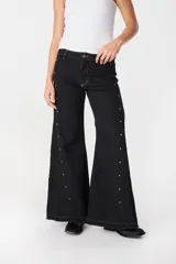 Pantalón de lino de pierna ancha, color negro, con diseño de godets laterales que aportan volumen y detalle de tachas metálicas doradas a lo largo de las costuras laterales. Presenta ruedo deshilachado.