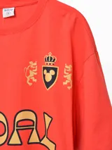 Remera naranja con cuello polo blanco, estampado frontal con la palabra "GOAL" en amarillo y negro, y logos bordados.