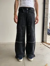 Pantalon baggy-flared de denim negro con efecto gastado, diseño desmontable con cierres horizontales en las piernas. Presenta múltiples parches de refuerzo, apliques metálicos, ojales decorativos y correas ajustables en los tobillos. Incluye un cinturón textil con tachas.