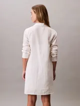 Vestido camisero corto de color blanco, confeccionado en tejido de gasa de algodón. Presenta cuello clásico, cierre frontal con botones, mangas largas y un bolsillo aplicado en el pecho.