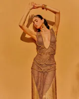 Vestido largo de tejido crochet en color bronce, con escote en V profundo, diseño de red y detalles trenzados. Presenta una silueta ajustada y un acabado con flecos largos en el ruedo.