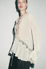 Blusa de escote en pico con manga larga terminada en volantes. Presenta aplicaciones de encaje a tono, cierre frontal con lazadas y bajo asimétrico.