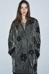 Kimono de terciopelo gris con acabado texturizado, cuello redondo y manga larga. Presenta bordados florales en hilo negro y metalizado, bolsillos delanteros y cierre frontal.