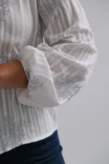 Blusa blanca de manga larga con diseño de rayas verticales y bordados florales en tono grisáceo. Presenta cuello camisero, cierre frontal con botones y puños elásticos.