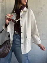 Chaqueta tipo sobrecamisa color beige, con cierre frontal de botones metálicos, cuello clásico y bolsillos frontales. Presenta un diseño de corte recto y mangas largas con puños abotonados.