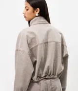 Campera bomber color gris de sarga, con cuello alto, cierre frontal con botones y bolsillos con solapa en el pecho. Presenta mangas largas con puños elásticos y ruedo ajustable con elástico.